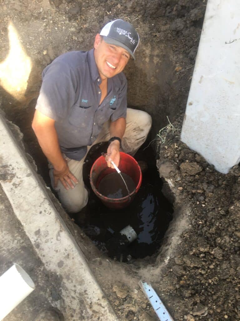 man with blue cap scooping water from a ground hole near pipes
